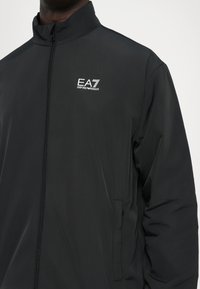 Black lightweight jacket featuring a high collar, front zipper, and EA7 logo on the chest. Smooth texture with a minimal design.