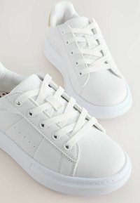 Adidași albi din piele sintetică texturată, cu șireturi plate, talpă din cauciuc și perforații subtile. Partea din spate prezintă un accent bej.