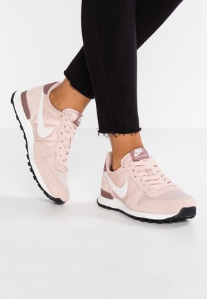 Person iført lyserøde Nike-sneakers med hvid swoosh og mauve accenter, kombineret med sorte bukser med frynset kant på hvidt gulv.
