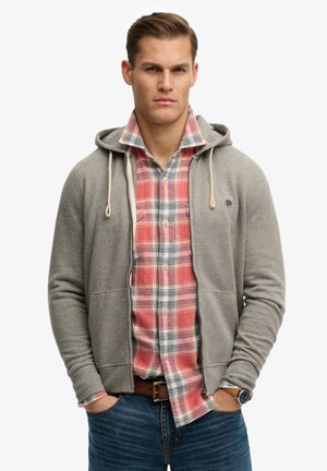 Mand iført en grå hættetrøje med lynlås over en rød- og blåtternet skjorte, blå jeans, brunt bælte og armbåndsur med hænderne i lommerne.