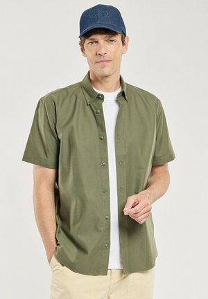 Homme portant une casquette bleue, une chemise olive à manches courtes boutonnée sur un t-shirt blanc, et un pantalon beige, debout avec une main dans la poche.