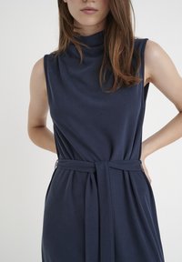 Granatowa sukienka bez rękawów z wysokim dekoltem, ściągnięta w pasie i z wiązanym paskiem. Miękka, zwiewna tkanina o bezszwowym, minimalistycznym wzorze.