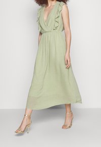 Vestido verde claro de longitud media con mangas rizadas y escote en V. Tela texturizada con rayas verticales. Lleva sandalias de tacón doradas.