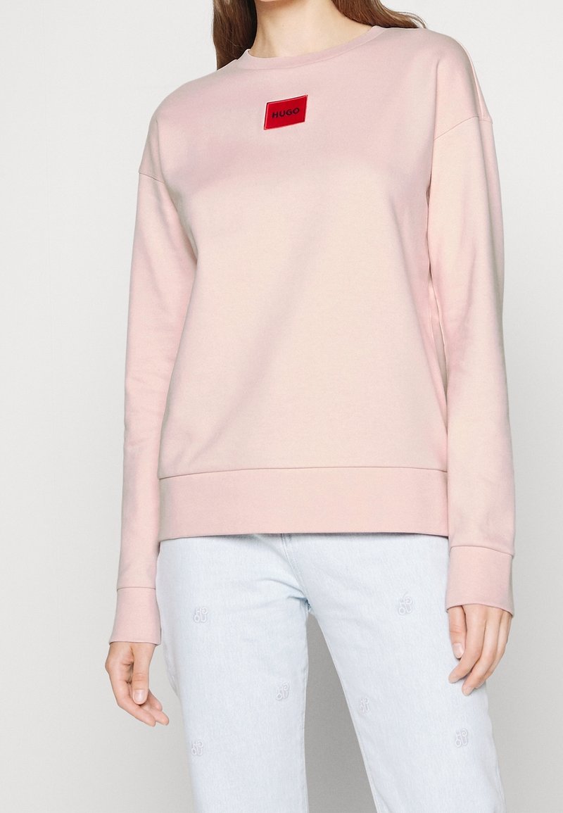 Felpa rosa chiaro con maniche lunghe, scollo rotondo e patch logo rosso. Tessuto morbido, orlo e polsini a costine. Abbinata a jeans azzurri.