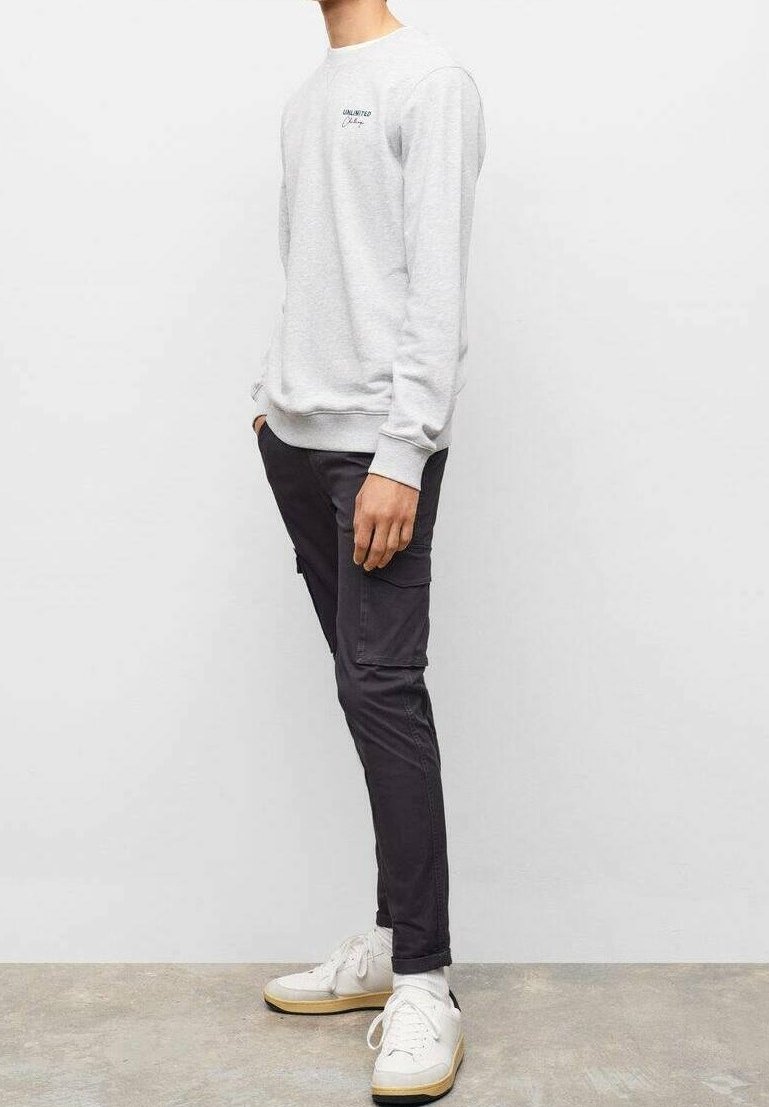 Homme debout de profil portant un sweat-shirt gris clair, un pantalon cargo noir, des chaussettes blanches et des baskets blanches sur un sol gris.
