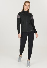 Schwarze, leichte Zip-Jacke mit grauen gemusterten Schulterakzenten, kombiniert mit schwarzen Leggings und grauen Sportschuhen. Definierte Passform und glatte Textur.