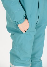 Teal vattentät overall med elastiska ärmmuddar, som har en sidoficka och "WX-PRO 10000" broderat på ärmen. Slät, hållbar textur.
