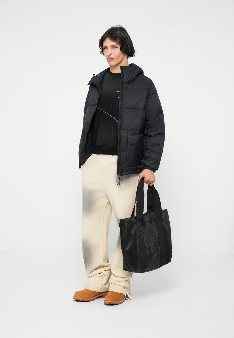 Μαύρο μπουφάν με κουκούλα, υφασμάτινη υφή και μπροστινή τσέπη; oversized παντελόνι σε ανοιχτό χρώμα με σκούρο gradient; μαύρη τσάντα τύπου tote με χερούλια.