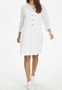 Weißes Augenlet-Kleid mit drei Viertel-Ärmeln, Knopf-Frontdetail, ausgestelltem Rock und strukturiertem Muster. Kombiniert mit weißen Sneakers.