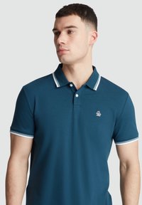 Donkerblauwe poloshirt met korte mouwen, voorzien van een witte gestreepte kraag en mouwboorden, met een klein logo op de linkerborst. Zacht materiaal.