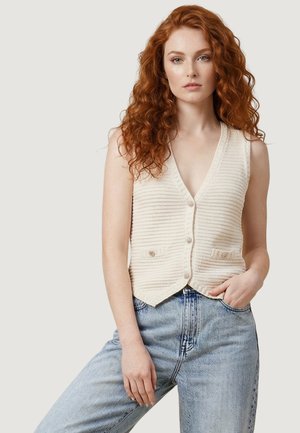 Giovane donna con lunghi capelli rossi ricci indossa un gilet di maglia color crema con bottoni e jeans a vita alta azzurri, in piedi davanti a uno sfondo semplice.