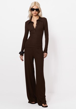 Femeie blondă purtând ochelari de soare negri supradimensionați, bluză lungă cu mâneci striate maro închis cu guler, pantaloni largi asortați și sandale negre.