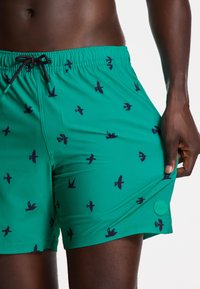 Green swim shorts featuring a black bird pattern, elastic waist with drawstring, and a logo patch on the hem. Made from smooth fabric.
