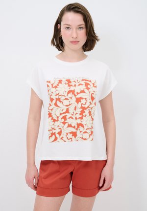 Weißes T-Shirt mit kurzen Ärmeln, das ein zentrales, orangefarbenes Blumenmuster mit cremefarbenen Details zeigt. Kombiniert mit rostfarbenen Shorts.