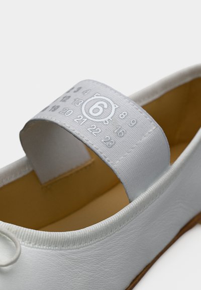 Λευκό δερμάτινο παπούτσι μπαλαρίνα με λαστιχένιο λουράκι που φέρει τον αριθμό 6 μέσα σε κύκλο και άλλους τυπωμένους αριθμούς.