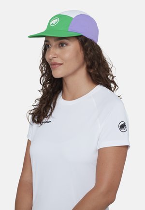 Donna con capelli ricci che indossa un berretto verde, viola e bianco e una maglietta bianca con i loghi Mammut sul petto e sulla manica.