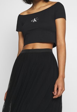 Kobieta ubrana w czarny dopasowany top odsłaniający ramiona z logo "CK Calvin Klein Jeans" oraz czarną teksturowaną spódnicę.