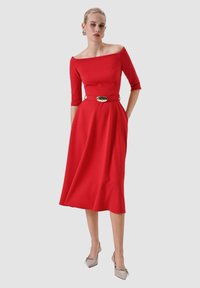 Abito rosso scollato con maniche a tre quarti, corpetto aderente e gonna svasata; cintura accentuata in oro e texture liscia.