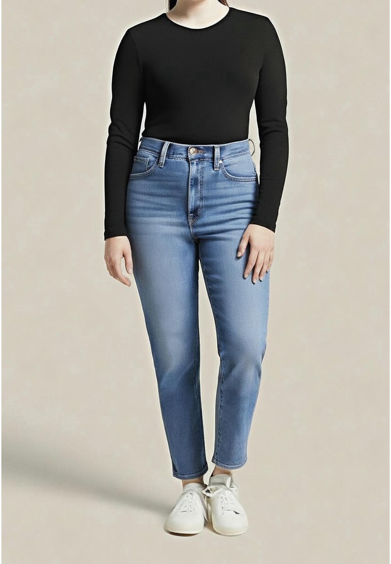 Personne portant un haut noir à manches longues, un jean bleu clair taille haute et des baskets blanches, debout devant un fond uni.