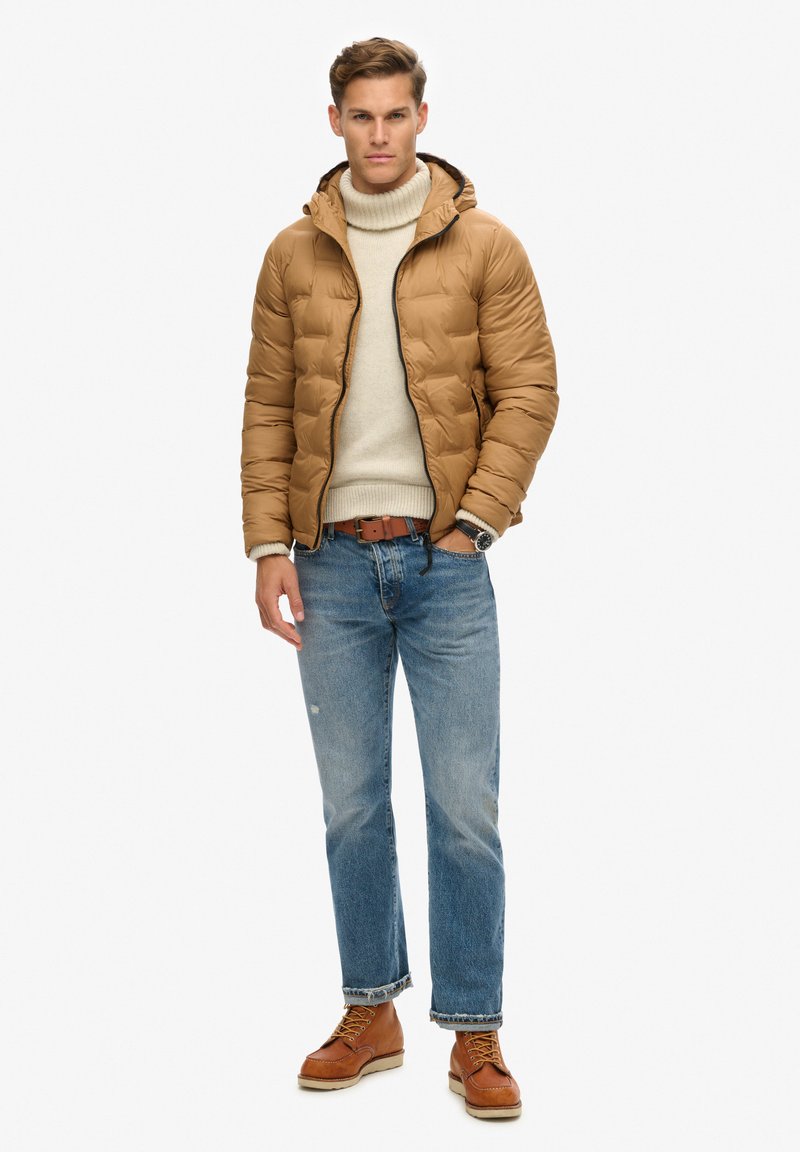 Tan pufferjack met capuchon, crèmekleurige coltrui, blauwe jeans en bruine veterschoenen. Opvallende details zijn ribgebreide manchetten en zoom.