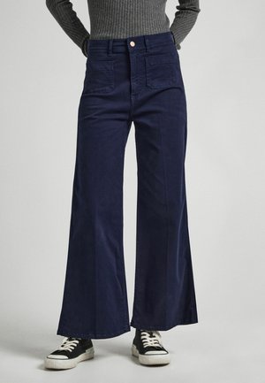Persona con pantalones anchos azul marino de talle alto y bolsillos delanteros, combinados con zapatillas negras y blancas y un suéter gris con textura acanalada.