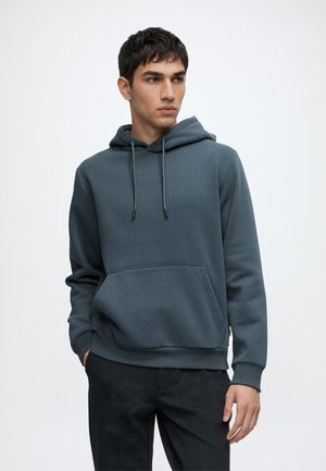 Jeune homme portant un sweat à capuche bleu sarcelle foncé avec poche avant et cordons, debout avec une main dans la poche regardant sur le côté.