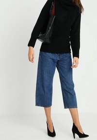 Schwarzer Rollkragenpullover, blaue zur Hüfte geschnittene Jeanshose und schwarze High-Heels. Schwarze glänzende Tragetasche mit roten Innen Details.