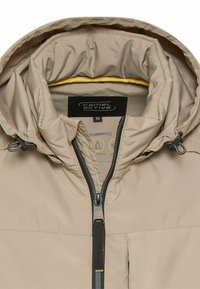 Beżowa kurtka outdoorowa z kapturem, z przodu zapinana na zamek, z regulowanymi sznurkami przy kapturze oraz widoczną metką marki wewnątrz kołnierza.