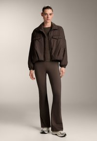 Brown puffer jacket with a collar and two front pockets, paired with fitted brown pants and light-colored sneakers. Smooth texture, relaxed fit.