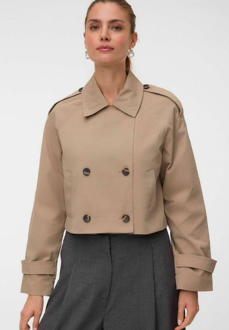 Femme portant une veste courte beige à double boutonnage avec un large col et un pantalon gris taille haute sur un fond uni.