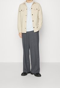 Beige textuur overhemdjasje met zwarte knopen, lichtblauwe sweatshirt eronder, grijze brede broek en zwarte boots met dikke zool.