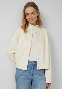 Cremefarbene, kurz geschnittene Jacke aus weichem Stoff; mit Druckknopfverschluss und geripptem Kragen. Kombiniert mit hellblauen Jeans.