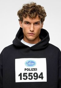Sweatshirt à capuche noir avec une grande étiquette blanche portant un texte bleu et noir, représentant "POLIZEI" et le numéro "15594."