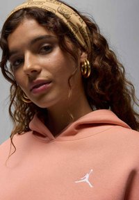 Sweat à capuche en tissu pêche doux, avec un logo Jumpman blanc. Cheveux coiffés en boucles lâches, soulignés par un bandeau à motif beige et des boucles d'oreilles en or.