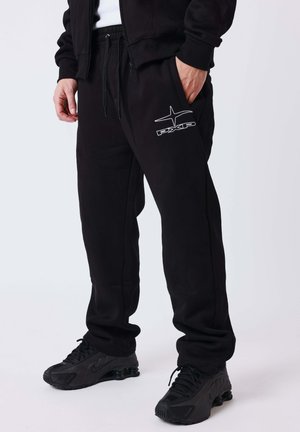 Man draagt zwarte joggingbroek met wit PXP-logo op de dij, zwarte sneakers en een zwart jack met rits, staand met één hand in de zak.