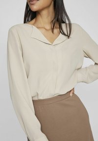 Donna che indossa una blusa beige a maniche lunghe infilata in una gonna marrone, con una mano poggiata sull'anca su uno sfondo neutro.
