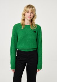 Groene gebreide trui met lange mouwen, geribbelde manchetjes, ronde hals en een klein zwart logo-detail op de borst. Gecombineerd met zwarte broeken.