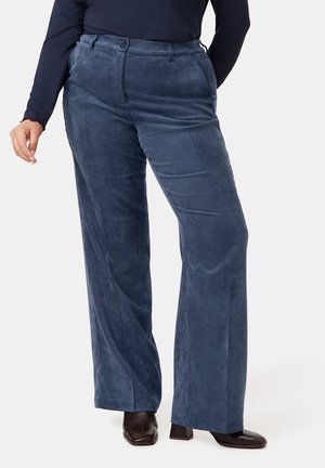 Osoba ubrana w niebieskie, szerokie spodnie sztruksowe z wysokim stanem i bocznymi kieszeniami, nosząca czarne buty na obcasie, stojąca na jednolitym tle.