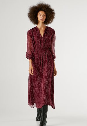 Robe midi bordeaux en tissu transparent avec des pois blancs, présentant un décolleté en V profond, une taille élastique et des manches à volants longues. Bottes noires jusqu'aux genoux.
