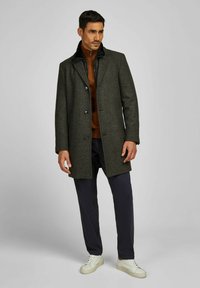 Donkergroen tweed overjas met een geknipt kraag, zwarte binnenvoering, twee knopen, gecombineerd met een bruine coltrui en marineblauwe broek.