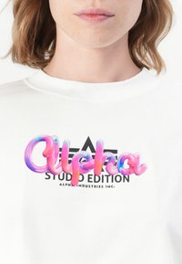 Detailný záber na biele tričko s farebným 3D textom "Alpha" a čiernym logom "Studio Edition Alpha Industries Inc." pod ním, ktoré nosí osoba.
