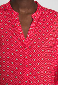 Blusa rossa con motivo geometrico in tonalità turchese e rosa, con scollatura a V, che mette in risalto una texture di tessuto leggero.