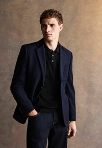 Marinblå blazer över en svart pikétröja, i kombination med mörka denimjeans. Har en strukturerad design, slät textur och subtila knappdetaljer.