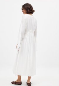 Hvid langærmet kjole med en indsnævret talje, flydende skørt og tekstureret stof. Har elegante ærmer og når anklerne.