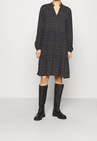 Černé květinové šaty s dlouhými rukávy, výstřihem do V a vrstvenou sukní. Nosič má na sobě černé vysoké boty s texturovaným povrchem.