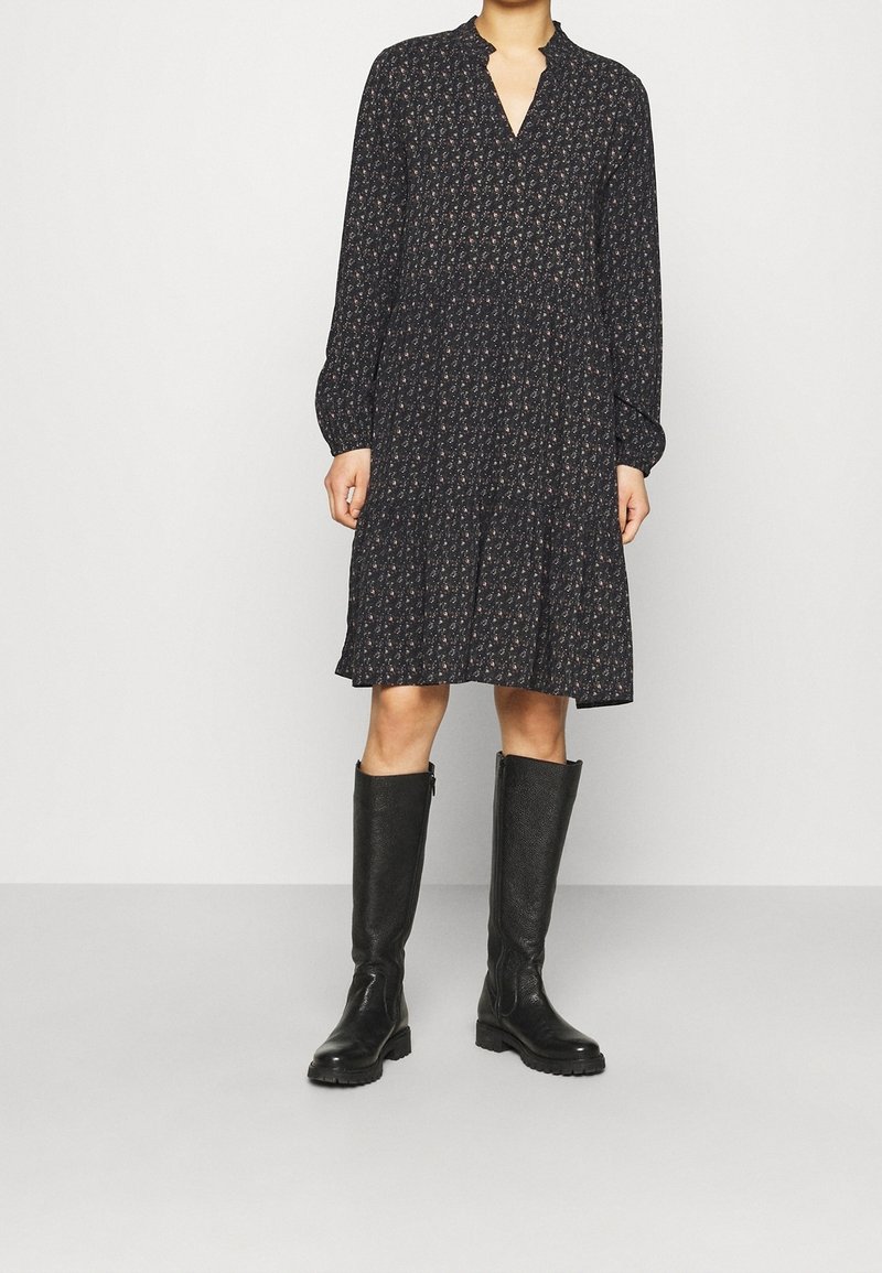 Černé květinové šaty s dlouhými rukávy, výstřihem do V a vrstvenou sukní. Nosič má na sobě černé vysoké boty s texturovaným povrchem.