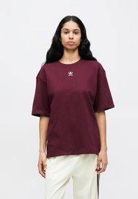 Burgundi pamut póló laza szabással, rövid ujjal és egy kis fehér Adidas logóval a mellkason. Világos színű nadrággal párosítva.