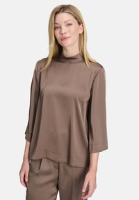Blusa marrone in satin con colletto alto, maniche a tre quarti e taglio A-line. Texture liscia con dettagli minimi e vestibilità rilassata.