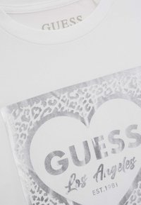 Fehér pamut póló, ezüst leopárd mintás szívvel, nagyméretű "GUESS" felirattal és alatta fekete "Los Angeles EST.1981" felirattal.