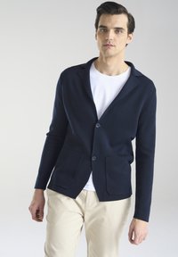 Granatowy kardigan z miękkiej dzianiny, z kołnierzem z wyłogami, dwiema przednimi kieszeniami i zapięciem na guziki, noszony na białej koszuli.